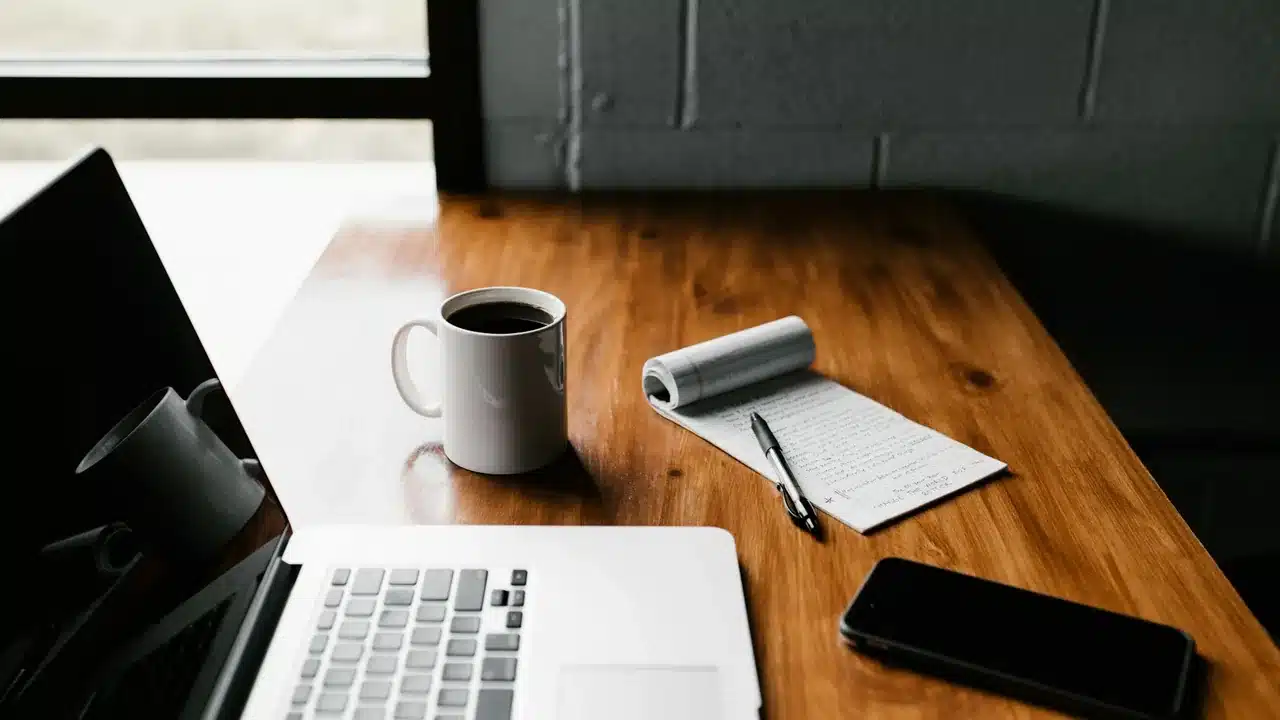 Bureau avec un ordinateur portable, un carnet de tâches et un café : les tâches répétitives à déléguer